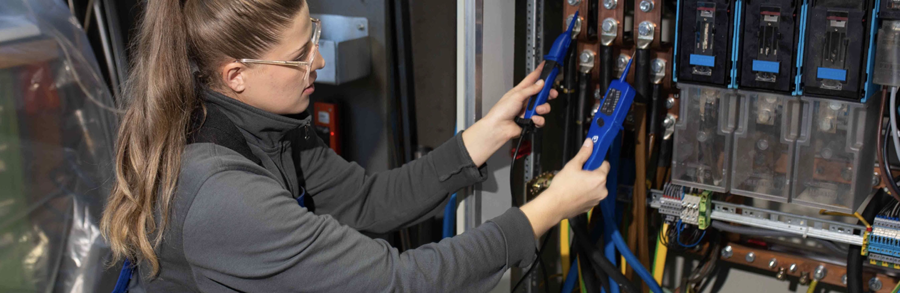 Weibliche Person arbeitet konzentriert mit einem Prüfgerät an einer elektrotechnischen Anlage. 
