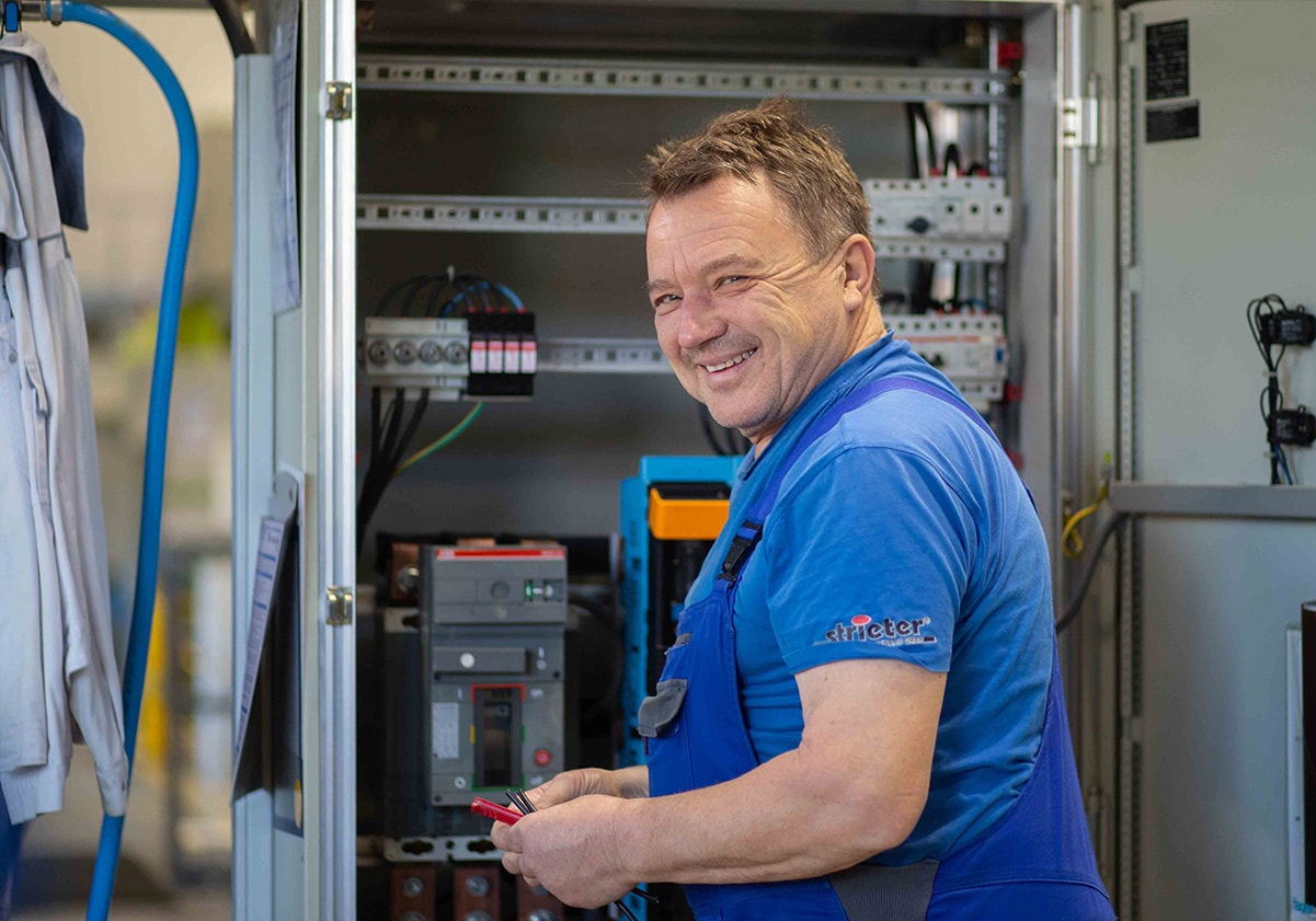 Strieter Mitarbeiter lächelt während der Arbeit an einem Schaltschrank in die Kamera