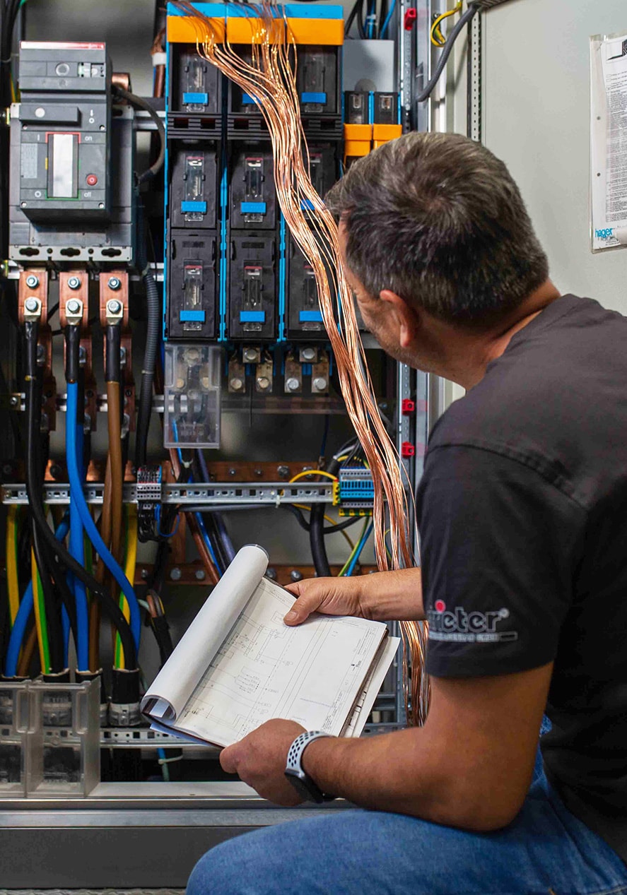 Ein Strieter Mitarbeiter blickt prüfend in einen offenen Schaltschrank und hat dabei den Plan einer elektrotechnischen Anlage in der Hand. 