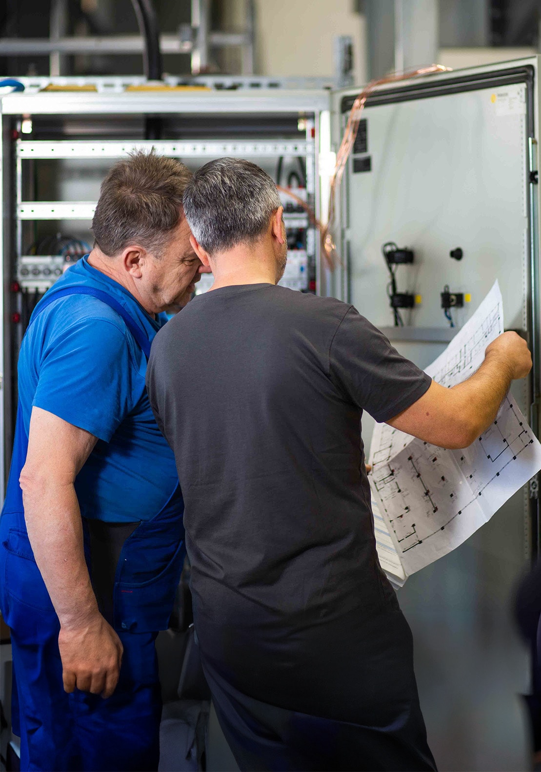 Zwei Kollegen besprechen den Plan einer elektrotechnischen Anlage während sie vor einem offenen Schaltschrank stehen.